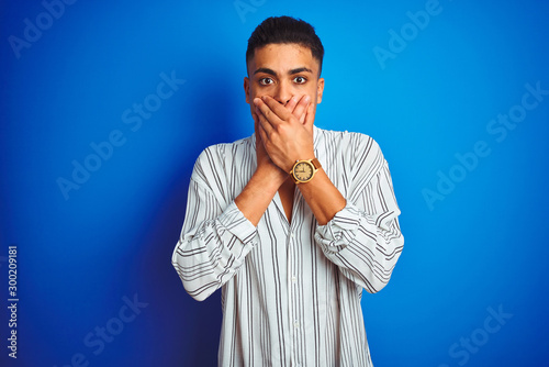 Young brazilian man wearing striped shirt standing over isolated blue background shocked covering mouth with hands for mistake. Secret concept.