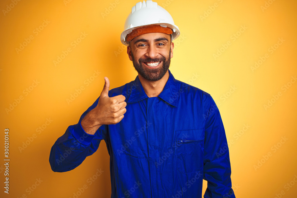 Handsome indian worker man wearing uniform and helmet over isolated yellow background doing happy thumbs up gesture with hand. Approving expression looking at the camera with showing success.