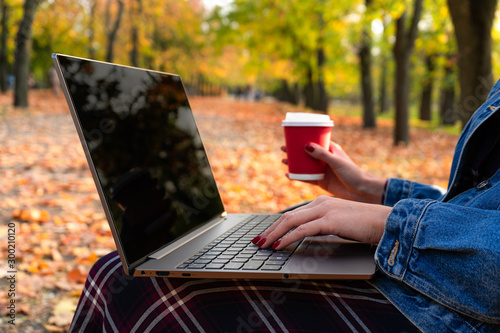 Girl in the autumn park on a bench with a laptop and coffee.