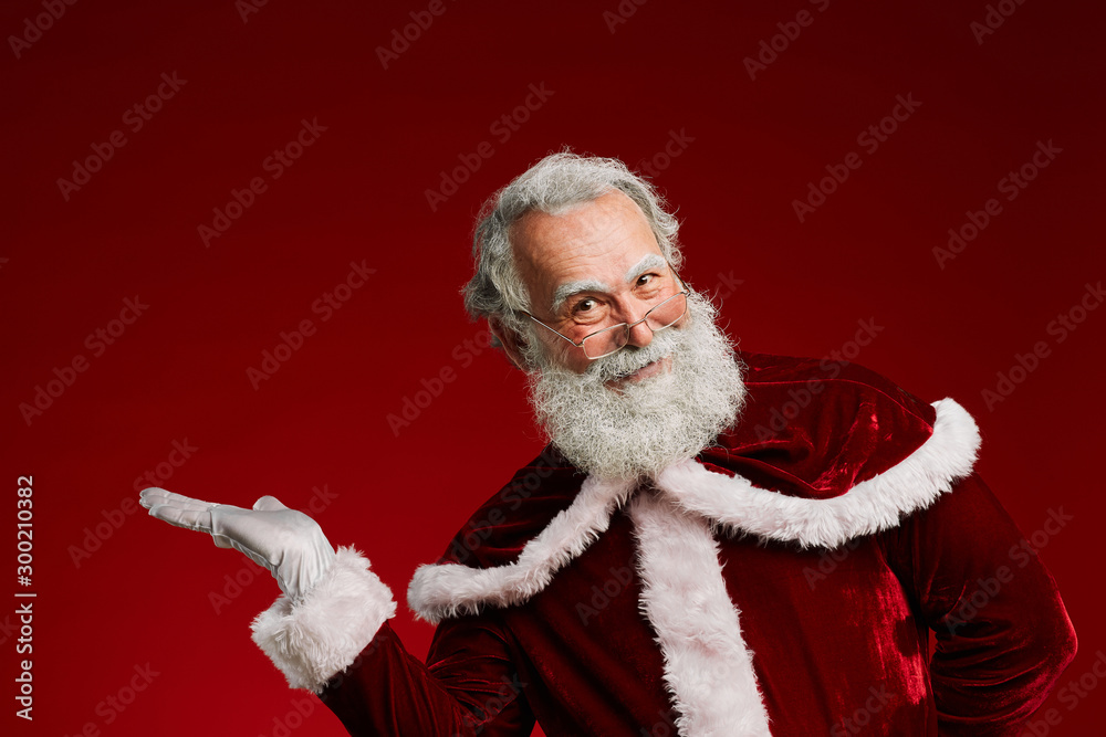 Naklejka premium Waist up portrait of cheerful Santa Claus looking at camera and pointing up while posing against red background, copy space