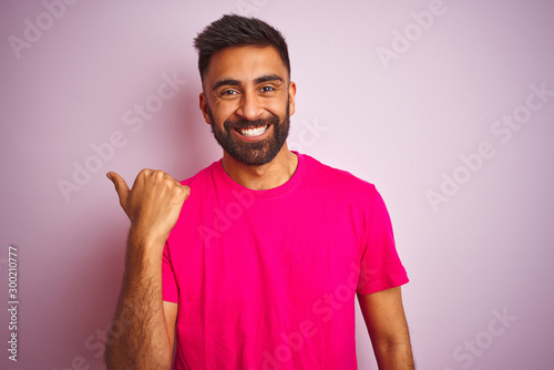 Young indian man wearing t-shirt standing over isolated pink background smiling with happy face looking and pointing to the side with thumb up.