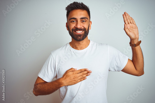 Young indian man wearing t-shirt standing over isolated white background smiling swearing with hand on chest and fingers up, making a loyalty promise oath photo