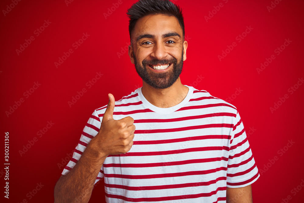 Young indian man wearing striped t-shirt standing over isolated red background doing happy thumbs up gesture with hand. Approving expression looking at the camera with showing success. Young indian man wearing striped t-shirt standing over isolated red background doing happy thumbs up gesture with hand. Approving expression looking at the camera with showing success.