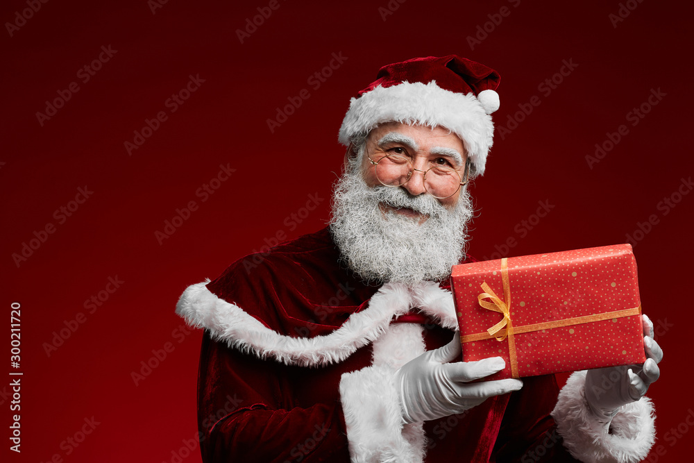 Naklejka premium Waist up portrait of classic Santa Claus holding Christmas present while standing against red background in studio, copy space