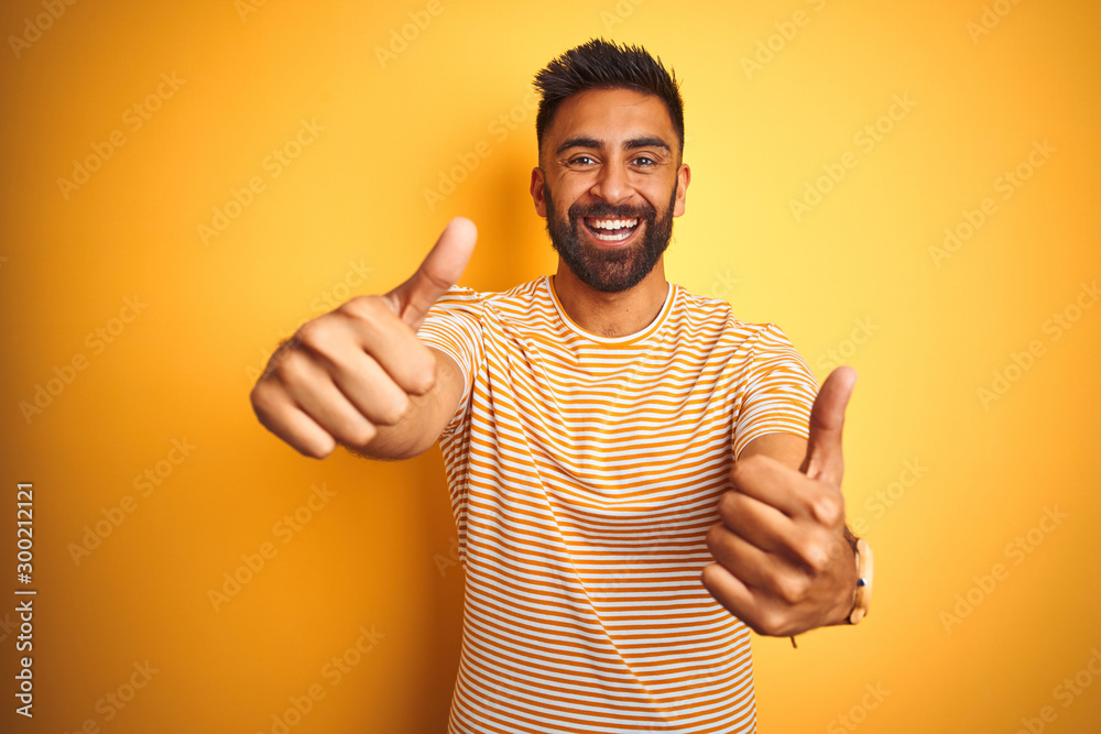 Φωτογραφία Young indian man wearing t-shirt standing over isolated yellow background approving doing positive gesture with hand, thumbs up smiling and happy for success