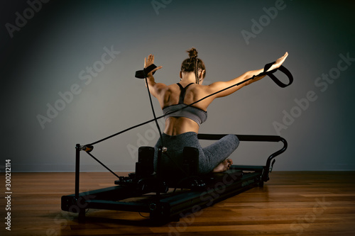 Young girl doing pilates exercises with a reformer bed. Beautiful slim fitness trainer on reformer gray background, low key, art light. Fitness concept