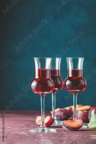 Plums strong alcoholic drink in grappas wineglass with dew. Hard liquor, slivovica, plum brandy or plum vodka with ripe plums on dark blue and  claret bordeaux concrete surface