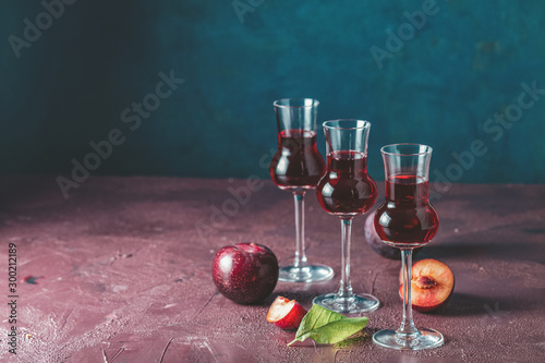 Plums strong alcoholic drink in grappas wineglass with dew. Hard liquor, slivovica, plum brandy or plum vodka with ripe plums on dark blue and  claret bordeaux concrete surface photo