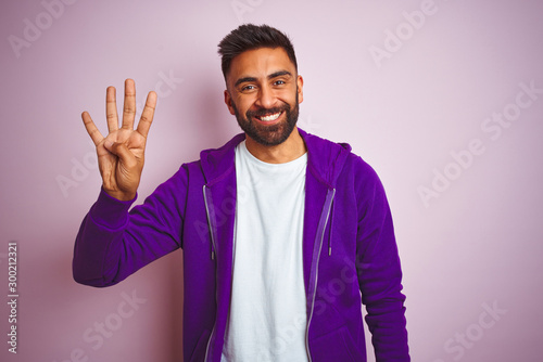 Young indian man wearing purple sweatshirt standing over isolated pink background showing and pointing up with fingers number four while smiling confident and happy. © Krakenimages.com