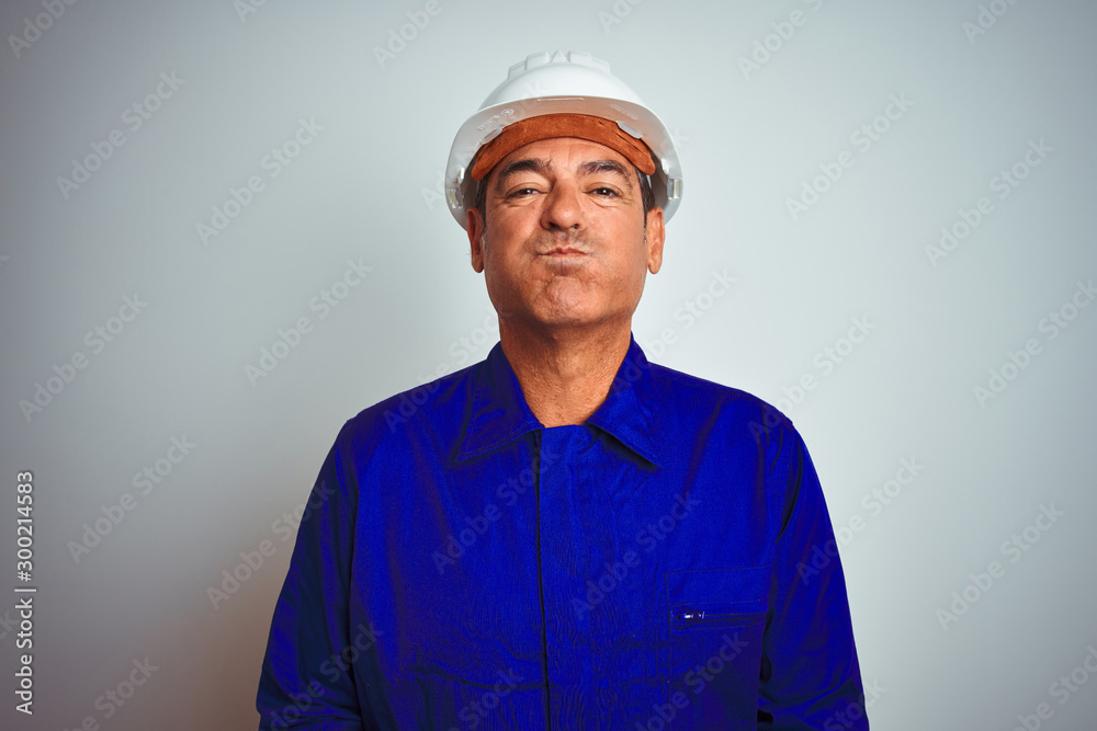 Handsome middle age worker man wearing uniform and helmet over isolated white background puffing cheeks with funny face. Mouth inflated with air, crazy expression.