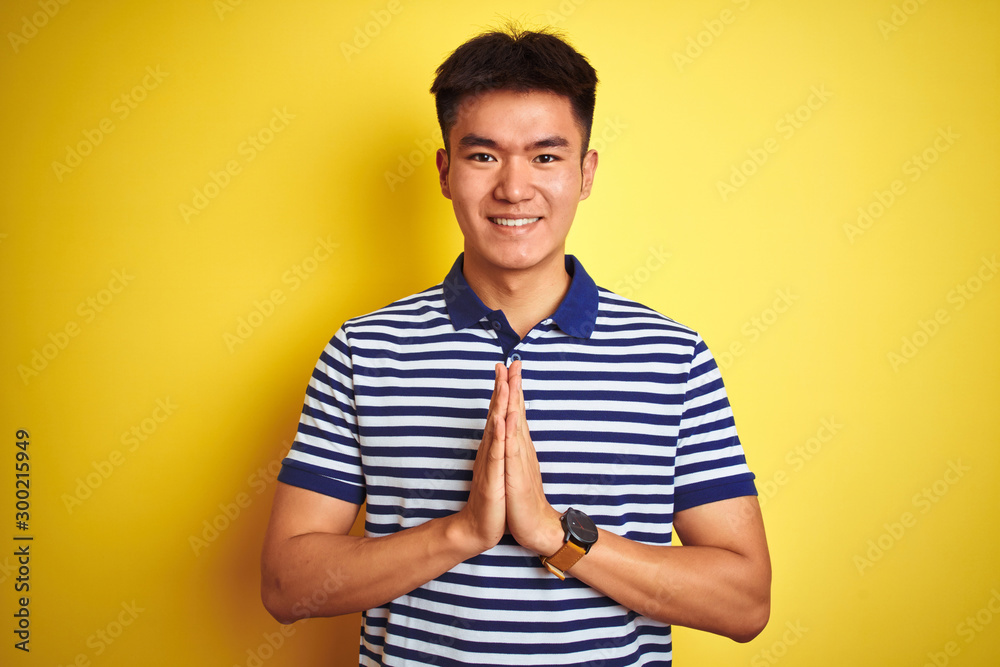 Young asian chinese man wearing striped polo standing over isolated yellow background praying with hands together asking for forgiveness smiling confident. Young asian chinese man wearing striped polo standing over isolated yellow background praying with hands together asking for forgiveness smiling confident.