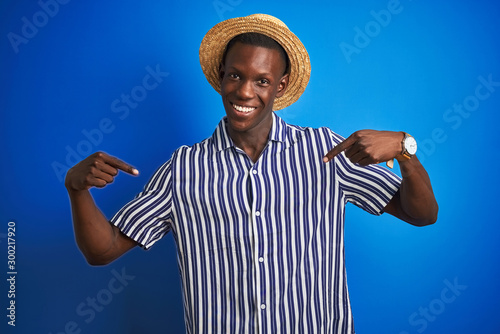 African american man wearing striped shirt and summer hat over isolated blue background looking confident with smile on face, pointing oneself with fingers proud and happy. © Krakenimages.com
