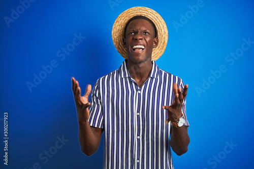 African american man wearing striped shirt and summer hat over isolated blue background crazy and mad shouting and yelling with aggressive expression and arms raised. Frustration concept. © Krakenimages.com