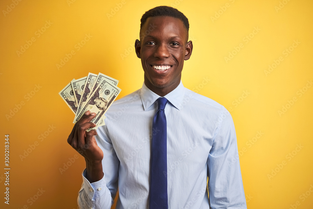 African american businessman holding dollars standing over isolated yellow background with a happy face standing and smiling with a confident smile showing teeth African american businessman holding dollars standing over isolated yellow background with a happy face standing and smiling with a confident smile showing teeth