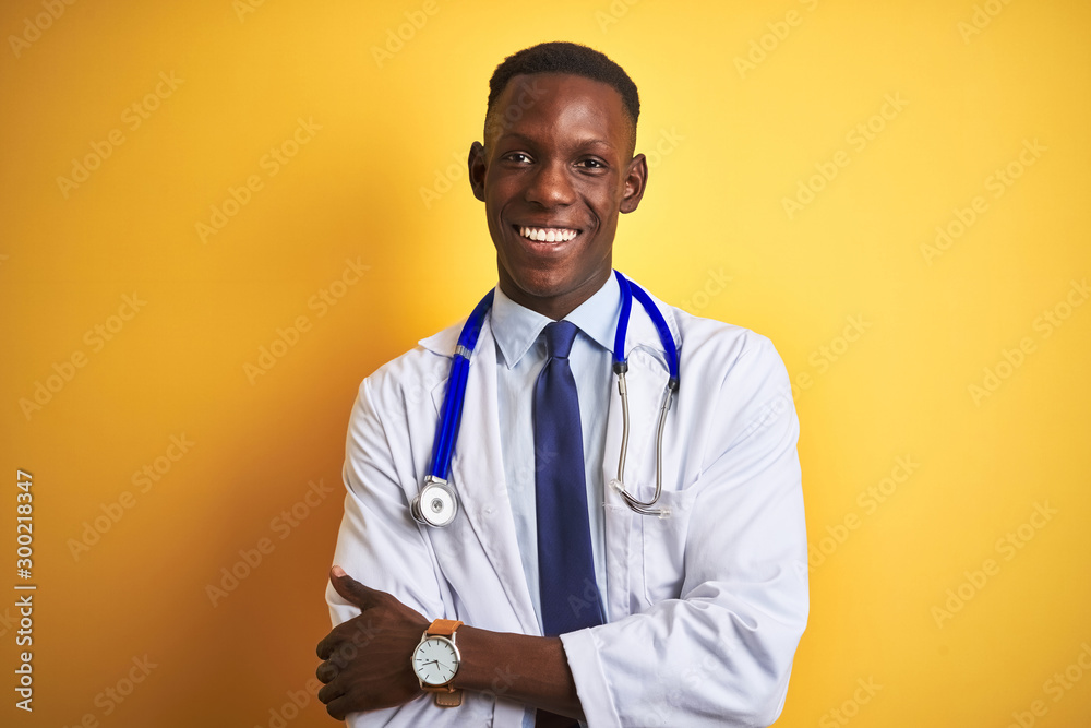 African american doctor man wearing stethoscope standing over isolated yellow background happy face smiling with crossed arms looking at the camera. Positive person. African american doctor man wearing stethoscope standing over isolated yellow background happy face smiling with crossed arms looking at the camera. Positive person.