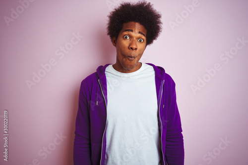 Young african american man wearing purple sweatshirt standing over isolated pink background making fish face with lips, crazy and comical gesture. Funny expression.