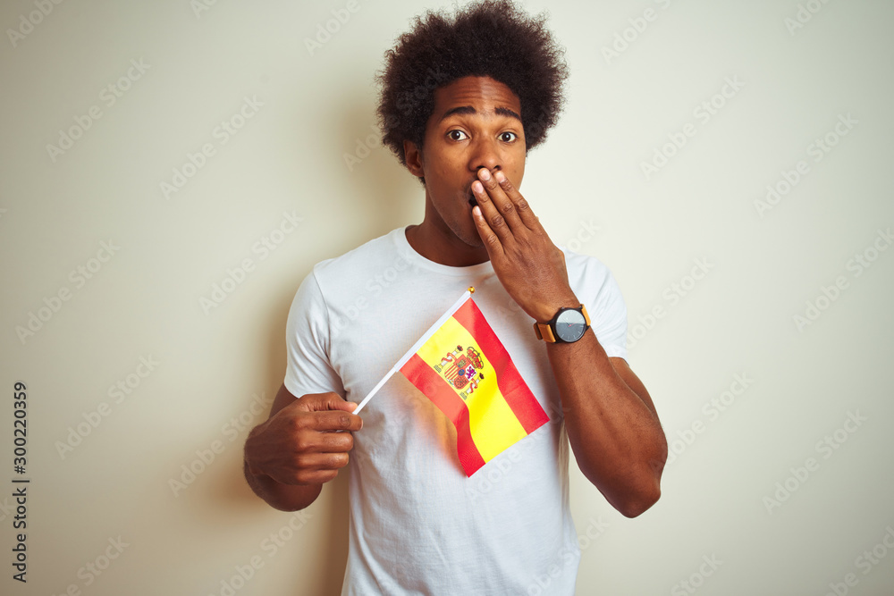 Young afro american man holding Spain Spanish flag standing over isolated white background cover mouth with hand shocked with shame for mistake, expression of fear, scared in silence, secret concept
