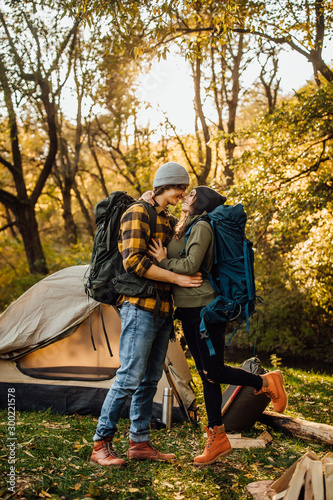 Young beautiful couple with hiking backpack kissing in the forest near tent. Attractive woman and handsome man relaxing together in nature. Instagram mood stories picture.