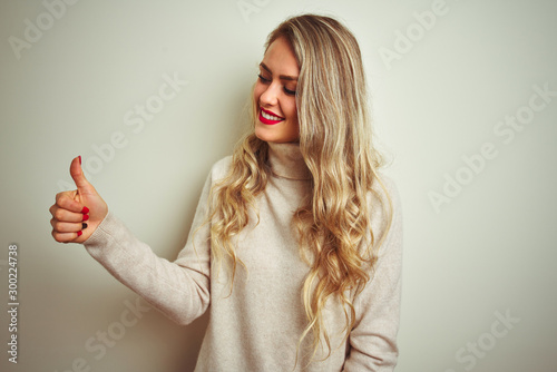 Beautiful woman wearing winter turtleneck sweater over isolated white background Looking proud, smiling doing thumbs up gesture to the side
