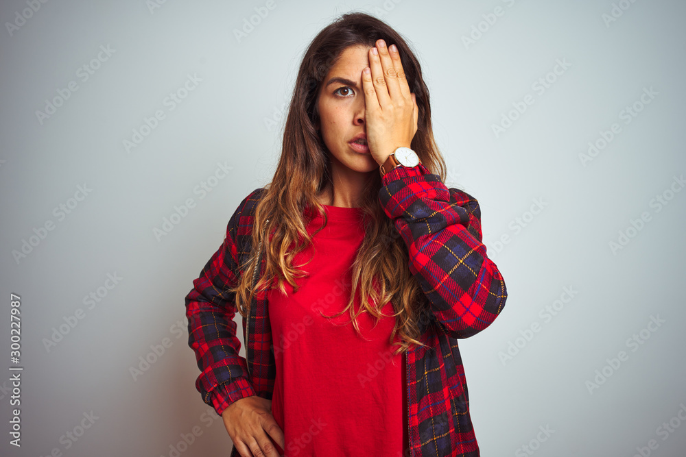 Young beautiful woman wearing red t-shirt and jacket standing over white isolated background covering one eye with hand, confident smile on face and surprise emotion. Young beautiful woman wearing red t-shirt and jacket standing over white isolated background covering one eye with hand, confident smile on face and surprise emotion.