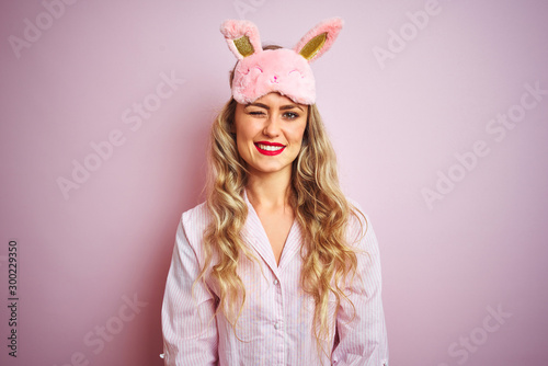 Young beautiful woman wearing pajama and sleep mask over pink isolated background winking looking at the camera with sexy expression, cheerful and happy face.