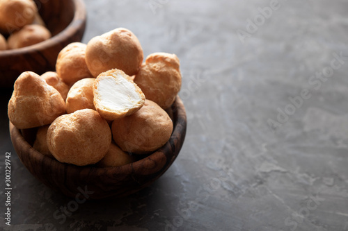 Small homemade round eclairs or profiteroles in a wooden bowl stand on a dark table. In the background is a wooden bowl with eclairs. Copy space