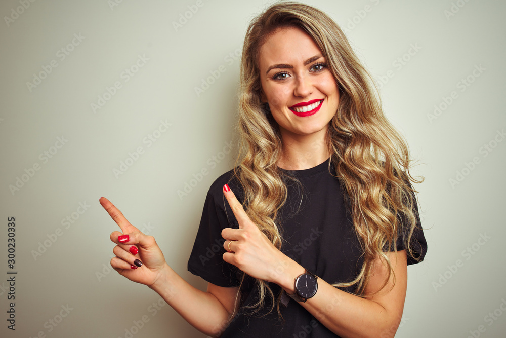 Young beautiful woman wearing black t-shirt standing over white isolated background smiling and looking at the camera pointing with two hands and fingers to the side. Young beautiful woman wearing black t-shirt standing over white isolated background smiling and looking at the camera pointing with two hands and fingers to the side.