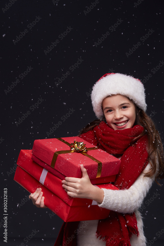 Young beautiful smiling girl in white knitting pullover, red scarf and Santa hat holding gift boxes on a dark background with snowflakes.Winter holidays, Christmas, New Year concept. Young beautiful smiling girl in white knitting pullover, red scarf and Santa hat holding gift boxes on a dark background with snowflakes.Winter holidays, Christmas, New Year concept.