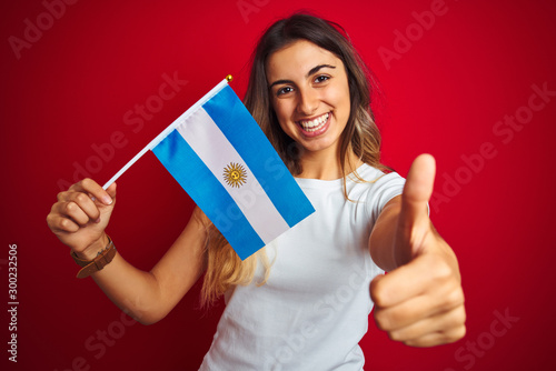 Young beautiful woman holding argentine flag over red isolated background happy with big smile doing ok sign, thumb up with fingers, excellent sign