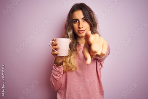 Young beautiful woman holding cup of coffee over pink isolated background pointing with finger to the camera and to you  hand sign  positive and confident gesture from the front