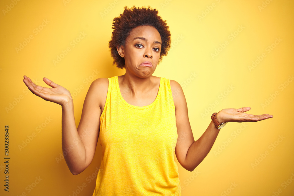 Beauitul african american woman wearing summer t-shirt over isolated yellow background clueless and confused expression with arms and hands raised. Doubt concept. Beauitul african american woman wearing summer t-shirt over isolated yellow background clueless and confused expression with arms and hands raised. Doubt concept.