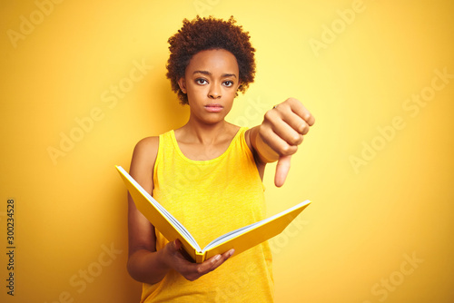 African american woman reading a book over yellow isolated background with angry face, negative sign showing dislike with thumbs down, rejection concept