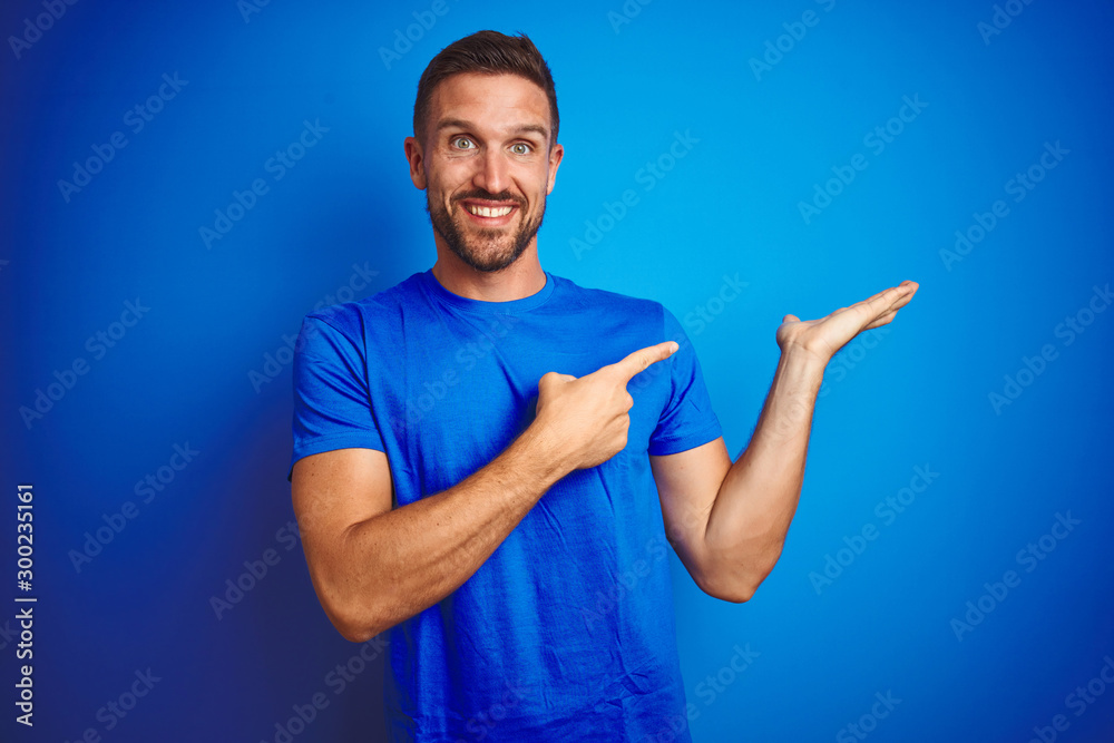 Young handsome man wearing casual t-shirt over blue isolated background amazed and smiling to the camera while presenting with hand and pointing with finger. Young handsome man wearing casual t-shirt over blue isolated background amazed and smiling to the camera while presenting with hand and pointing with finger.