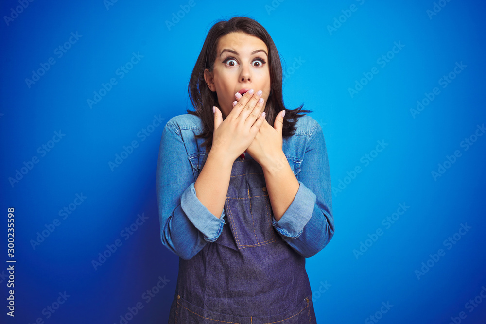 Young beautiful business woman wearing store uniform apron over blue isolated background shocked covering mouth with hands for mistake. Secret concept. Young beautiful business woman wearing store uniform apron over blue isolated background shocked covering mouth with hands for mistake. Secret concept.
