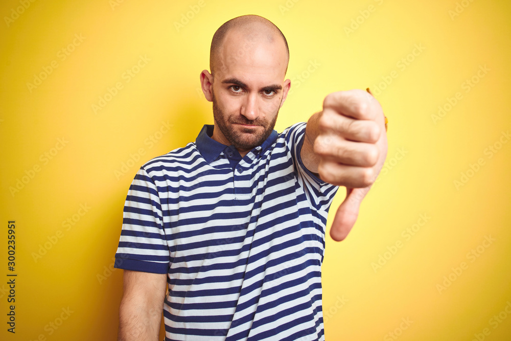 Naklejka premium Young bald man with beard wearing casual striped blue t-shirt over yellow isolated background looking unhappy and angry showing rejection and negative with thumbs down gesture. Bad expression.