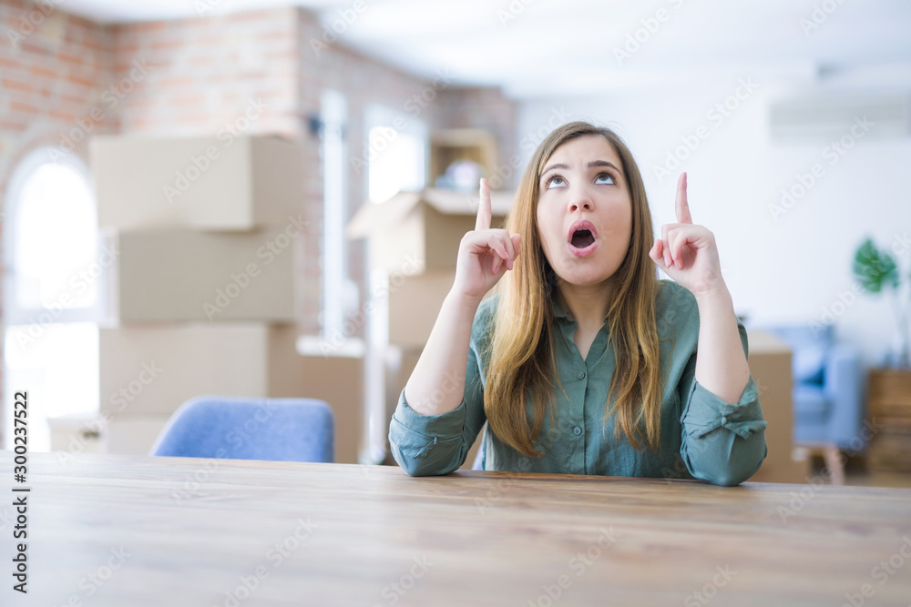 Young woman sitting on the table with cardboard boxes behind her moving to new home amazed and surprised looking up and pointing with fingers and raised arms.