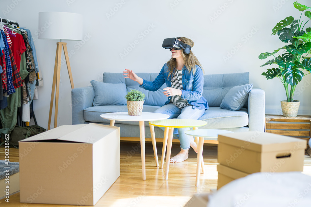 Young blonde woman wearing virtual reality glasses playing a simulation game sitting on the sofa around cardboard boxes moving to a new house Young blonde woman wearing virtual reality glasses playing a simulation game sitting on the sofa around cardboard boxes moving to a new house