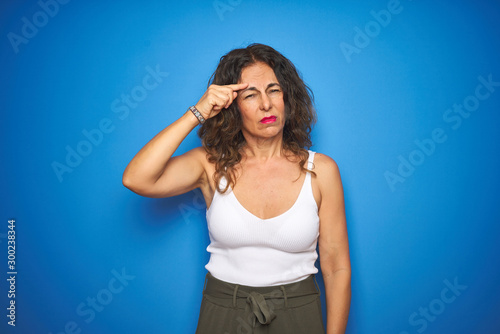 Middle age senior woman with curly hair standing over blue isolated background pointing unhappy to pimple on forehead, ugly infection of blackhead. Acne and skin problem
