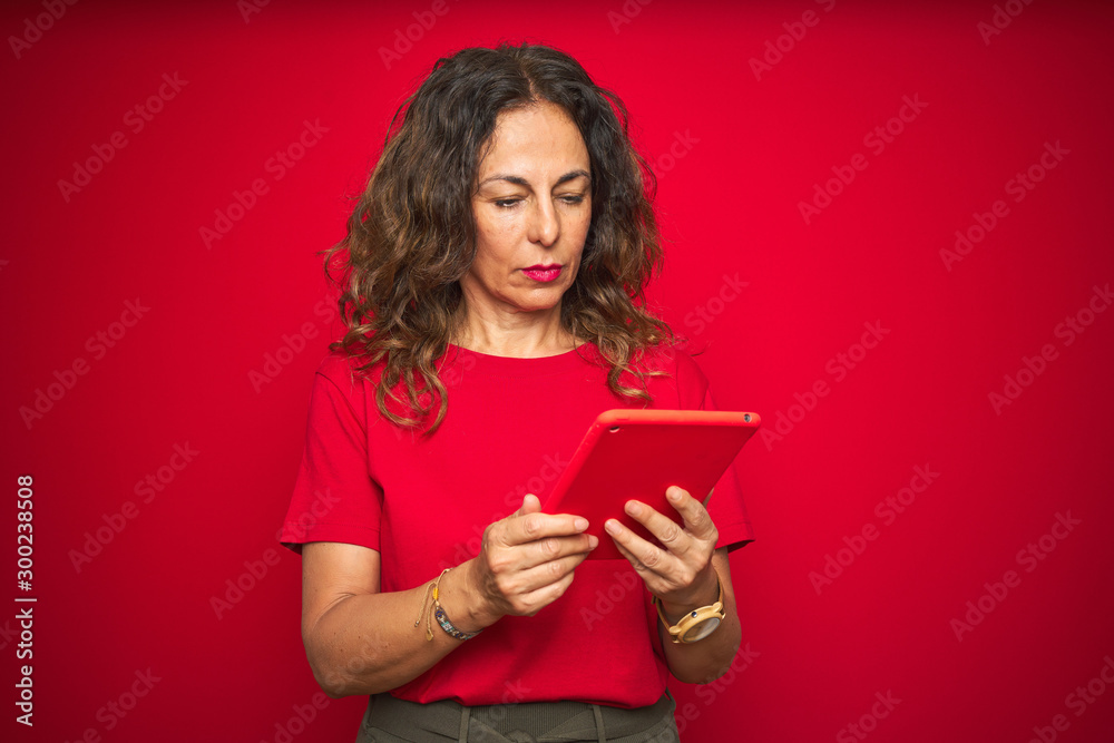 Middle age senior woman using touchpad tablet over red isolated background with a confident expression on smart face thinking serious Middle age senior woman using touchpad tablet over red isolated background with a confident expression on smart face thinking serious