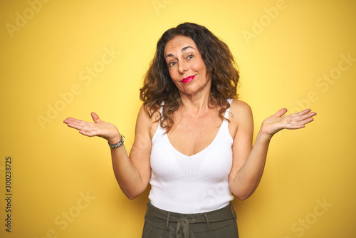 Middle age senior woman with curly hair standing over yellow isolated background clueless and confused expression with arms and hands raised. Doubt concept.