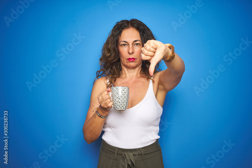 Middle age senior woman drinking a cup of coffee over blue isolated background with angry face, negative sign showing dislike with thumbs down, rejection concept