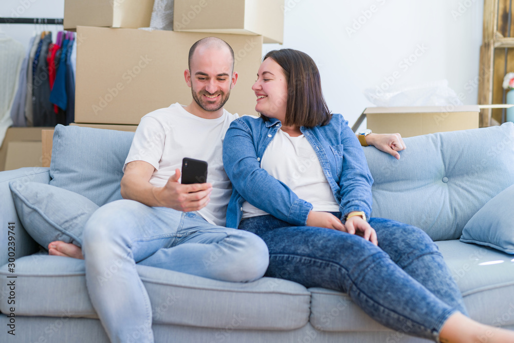 Young couple sitting on the sofa at new home using smartphone, hugging and smiling happy for moving to a new apartment