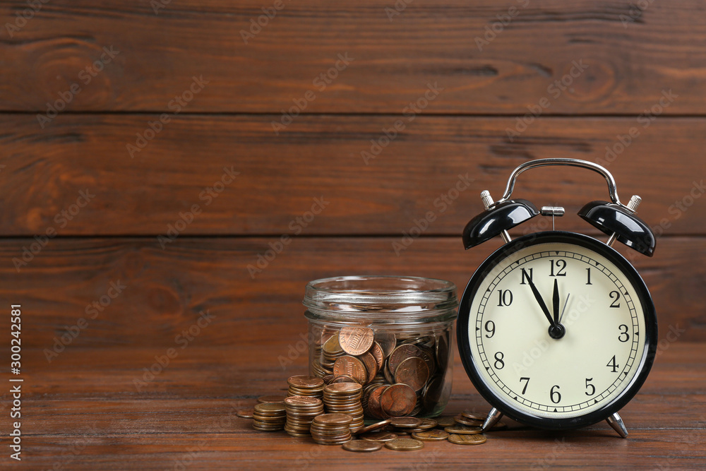 Black alarm clock, glass jar and coins on table against wooden background, space for text. Money savings Black alarm clock, glass jar and coins on table against wooden background, space for text. Money savings