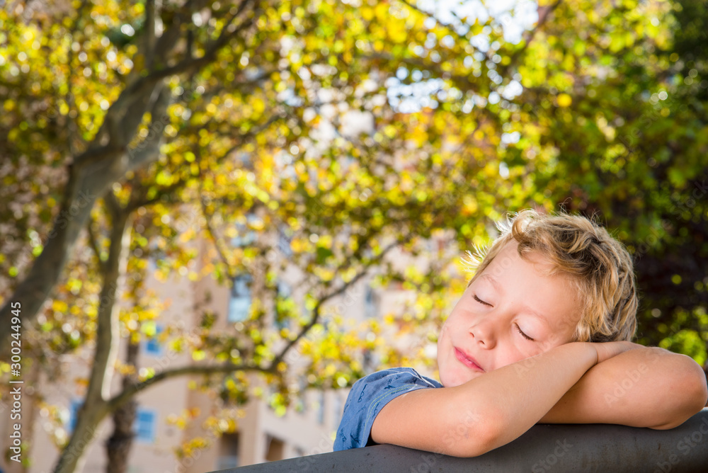 Obraz premium Portrait of blond boy with eyes closed calmly while resting with tree leaves background.