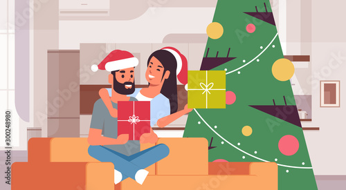 couple holding gift boxes merry christmas happy new year holiday celebration concept man woman embracing wearing santa hats sitting on couch near fit tree modern living room interior horizontal vector