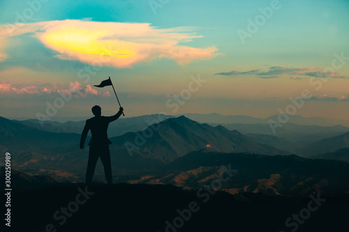 Silhouette of a man standing with a victory flag on the top of the mountain on sunrise sky background,business, success, leadership and achievement concept
