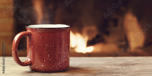 Red mug with hot tea in front of a burning fireplace, comfort, winter holidays and warmth of the hearth concept
