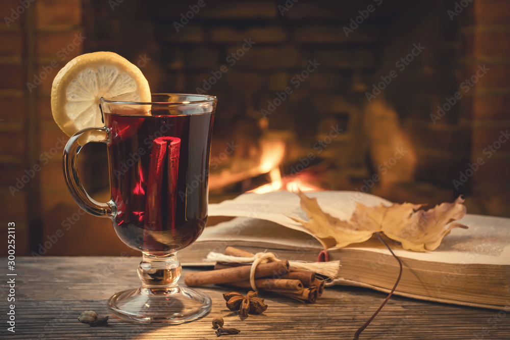 Glass of mulled wine, spices, a book on a table in a cozy room with a burning fireplace - obrazy, fototapety, plakaty 