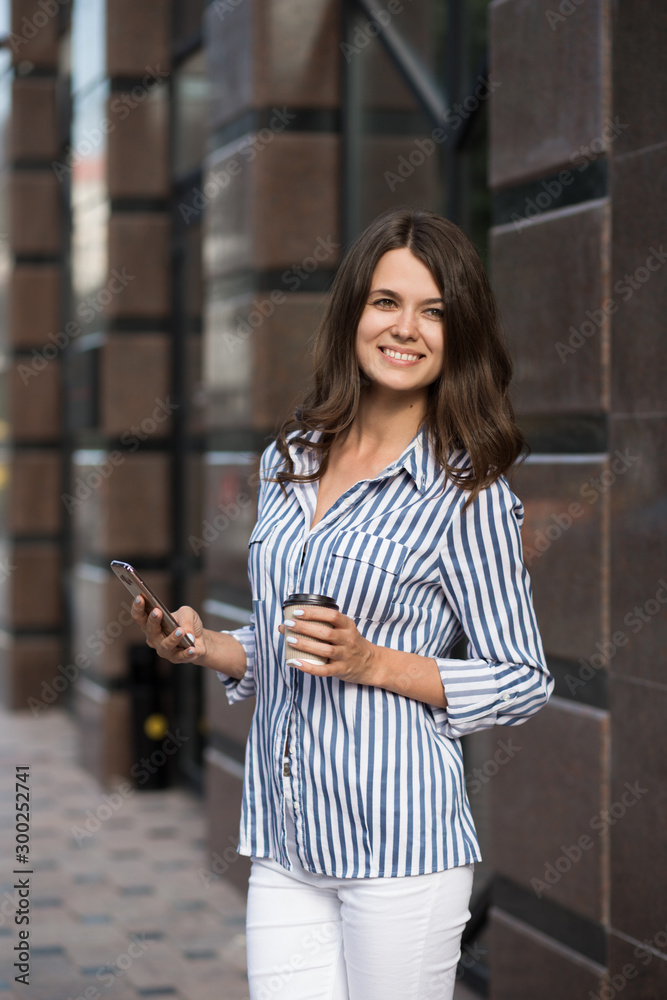 Businesswoman uses a mobile phone, walking along the city streets with coffee in her hand.