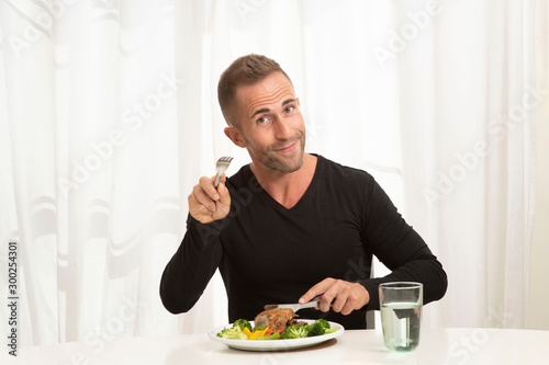 Handsome caucasian male wearing black casual top sitting at the dinning table eating a healthy meal of meat and vegetables and a glass of water pointing his fork at the camera